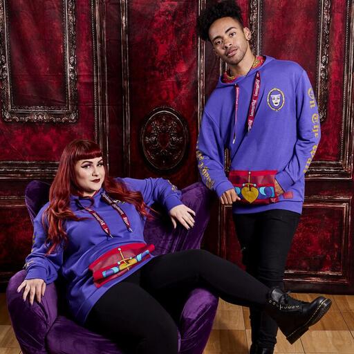 Woman lounging in a purple chair with a man standing beside her. Both are wearing the purple Unisex Heart Box Hoodie